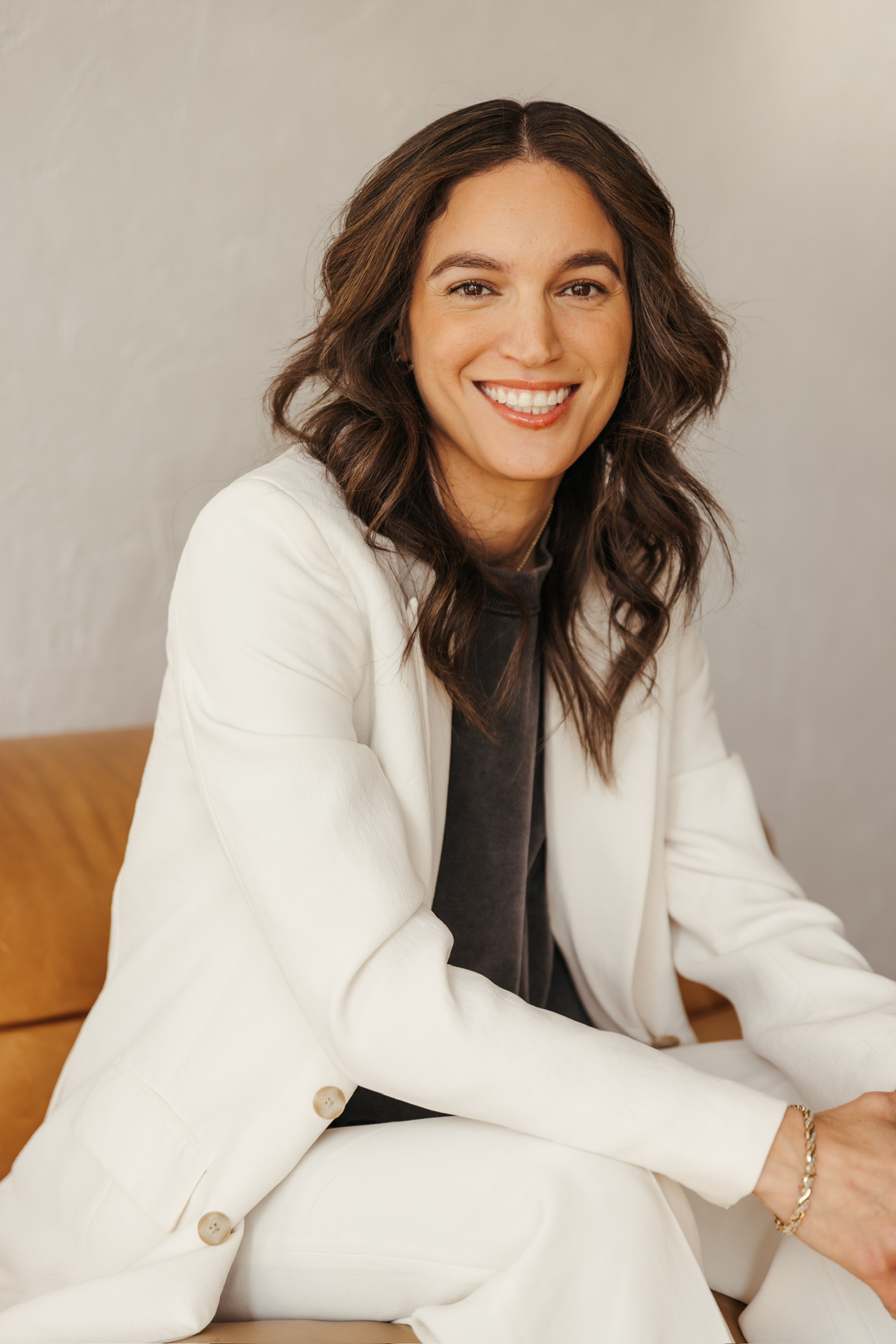 A photo of Alina Hernandez wearing a white suit, dark grey shirt, and gold bracelet