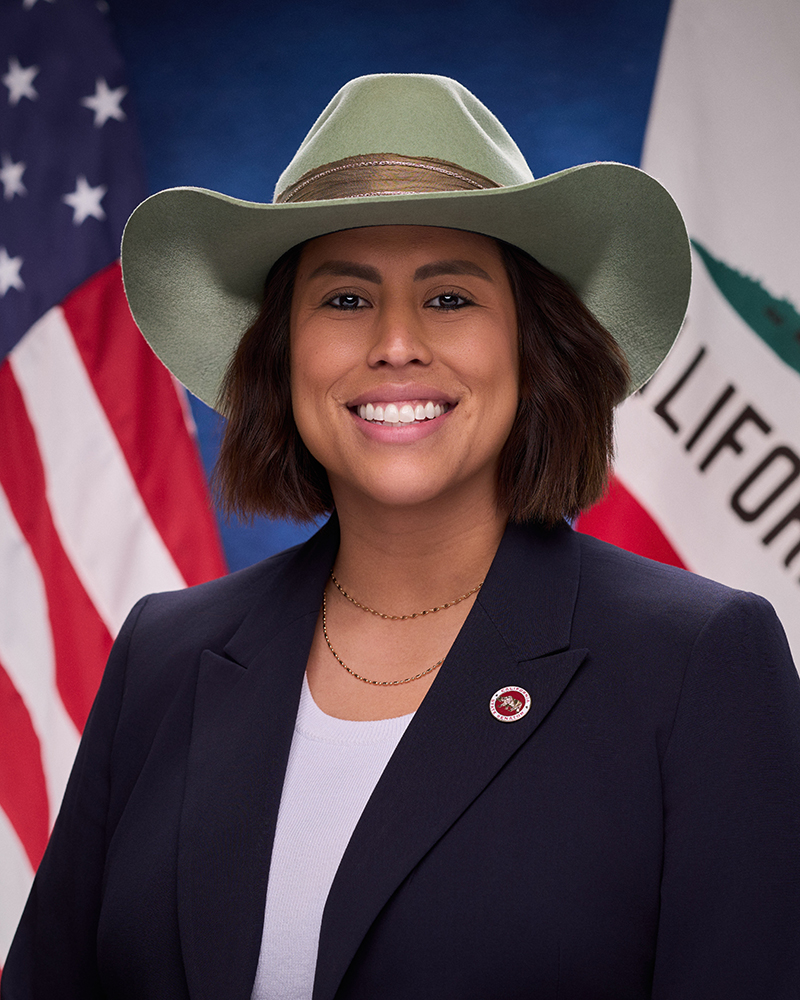 A photo of Senator Caroline Menjivar wearing a navy blue blazer, white shirt, gold chain necklace, and pistachio green cowboy hat